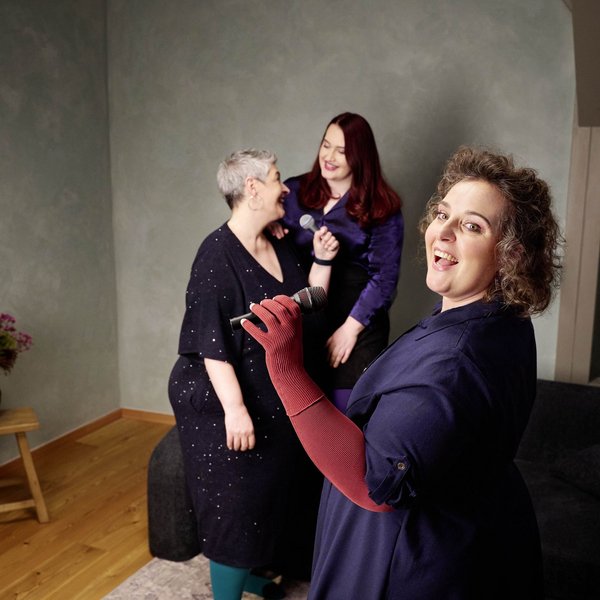 Drei Frauen stehen in einem Wohnzimmer mit hellgrüner Wand und singen gemeinsam mit Mikrofonen. Eine Frau im Vordergrund trägt dunkelblaues Kleid und rote Kompressionshandschuhe, während sie lächelnd in die Kamera singt. Im Hintergrund lachen zwei weitere Frauen, eine mit kurzen grauen Haaren und eine mit langen roten Haaren. Links im Bild steht ein Hocker mit einer Blumenvase.